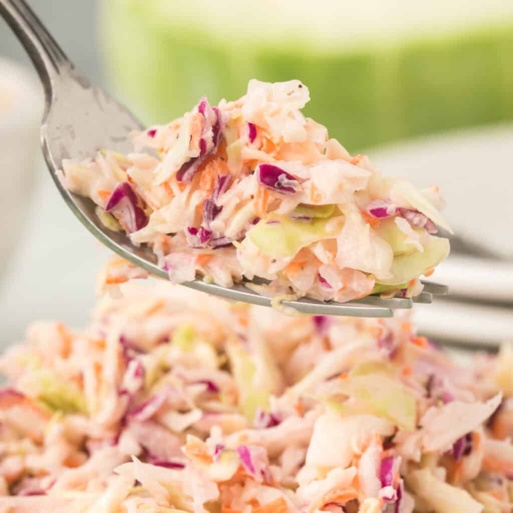 Square close-up of a forkful of creamy coleslaw lifted above a full bowl of salad.
