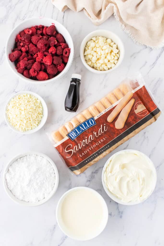 Ingredients for White Chocolate Raspberry Tiramisu laid out on a marble counter, including raspberries, white chocolate chips, powdered sugar, mascarpone, heavy cream, imitation rum, and ladyfingers.