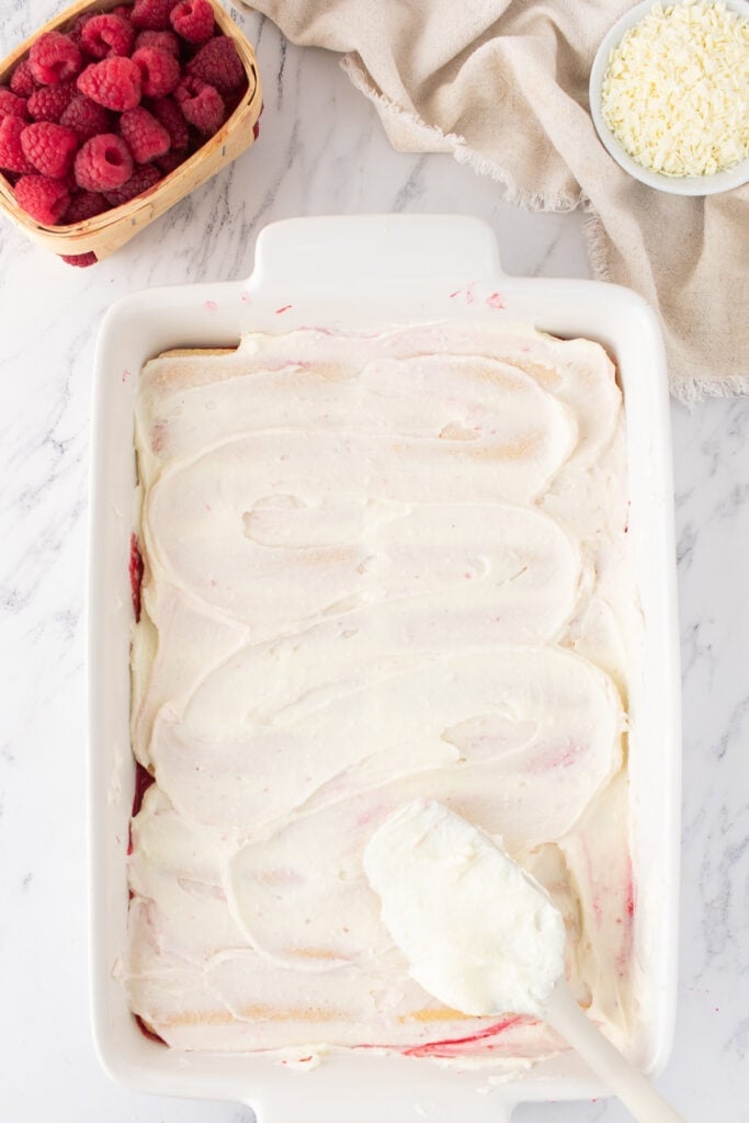 First layer of mascarpone cream spread evenly over ladyfingers in a baking dish, raspberries and grated chocolate in the background.