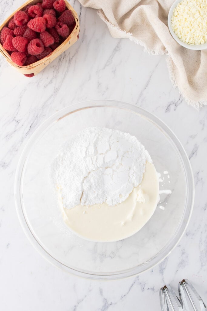 Bowl with powdered sugar and cream in the early stage of making mascarpone cream for tiramisu, fresh raspberries and white chocolate nearby.