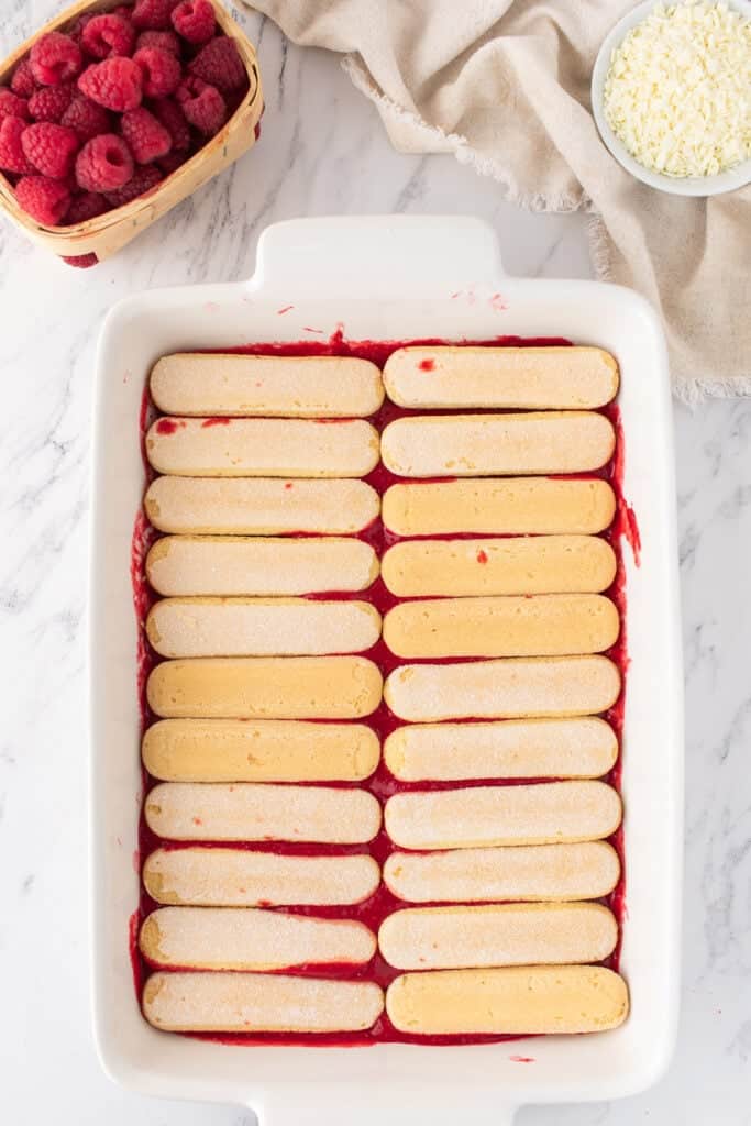 Ladyfinger biscuits arranged in a single layer over raspberry sauce in a white baking dish.
