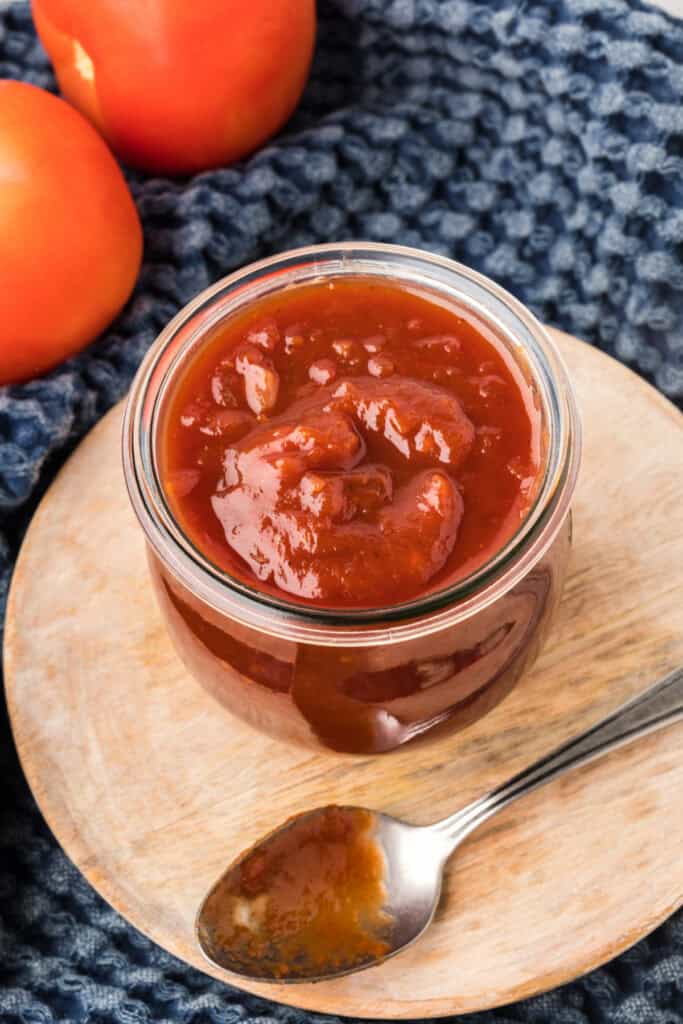 Jar of Bourbon Whiskey BBQ Sauce on a wooden board with a spoon beside it and tomatoes in the background