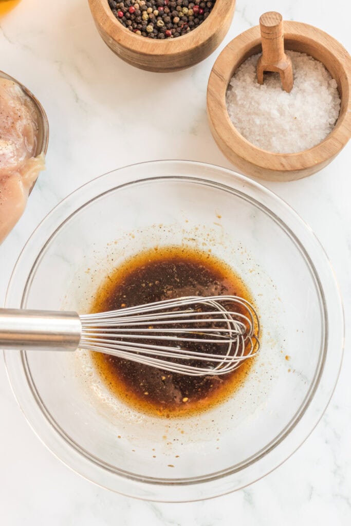 Whisk mixing marinade in a glass bowl next to peppercorns and salt