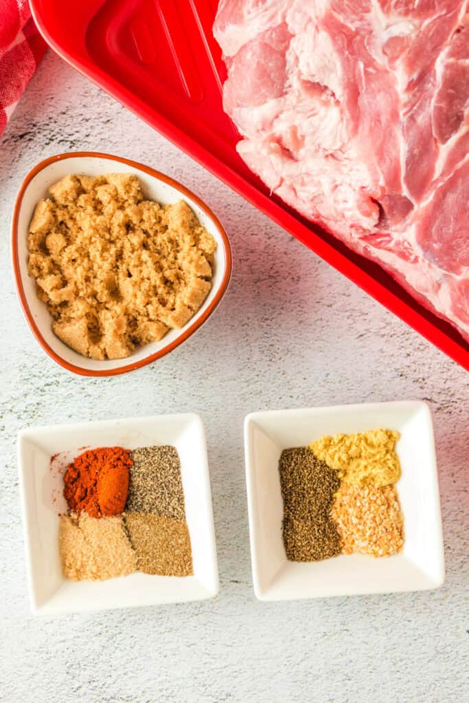 Overhead view of raw Boston butt in a red tray with separate spices and brown sugar ready for rub.