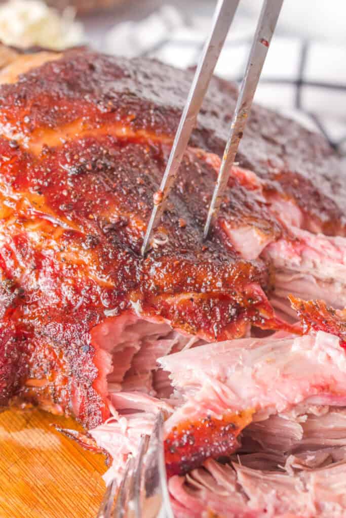 Forks shredding into tender, juicy grilled pulled pork on a cutting board.
