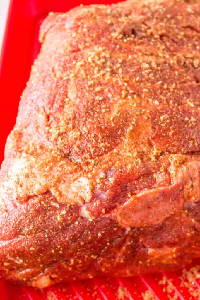 Raw Boston butt covered generously in dry rub, resting in a red tray.