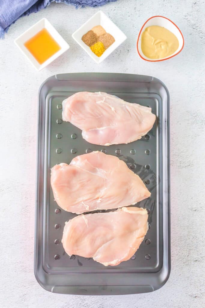 Raw chicken breasts on a marinade tray with seasonings, honey, and mustard nearby