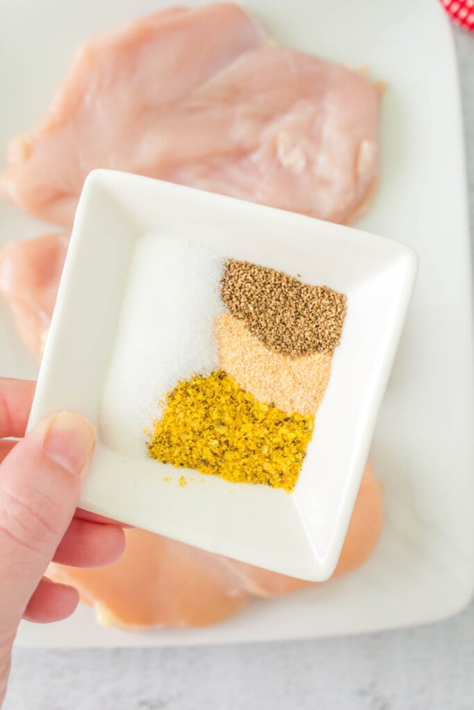 Small white dish with neatly arranged lemon pepper, garlic powder, salt, and celery seed held over raw chicken breasts.