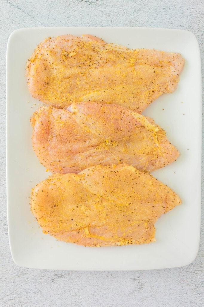 Raw chicken breasts coated with lemon pepper seasoning on a white plate.