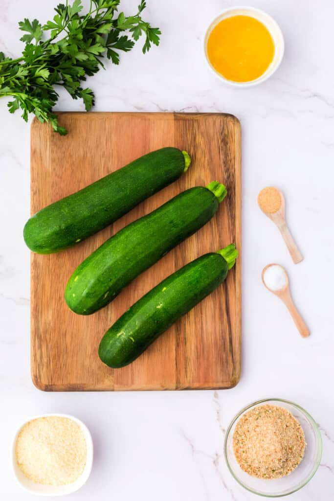 Whole zucchinis on a wooden cutting board with parsley, melted butter, garlic powder, salt, parmesan, and breadcrumbs.