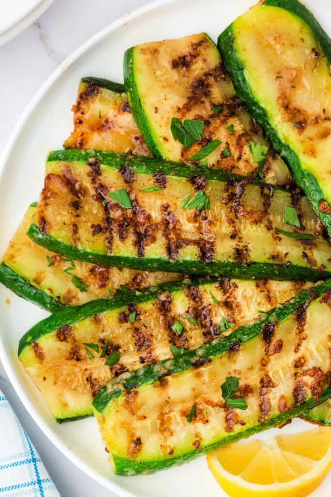 Grilled zucchini slices on a plate garnished with chopped parsley and a lemon wedge.