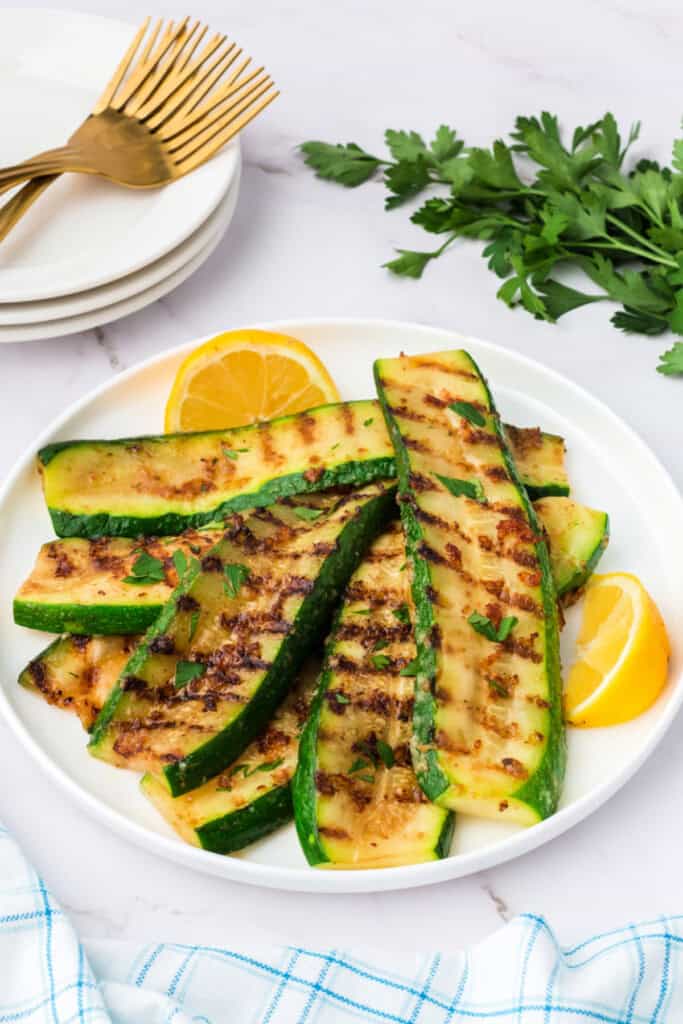 Side angle of grilled parmesan zucchini served on a white plate with fresh parsley and lemon.
