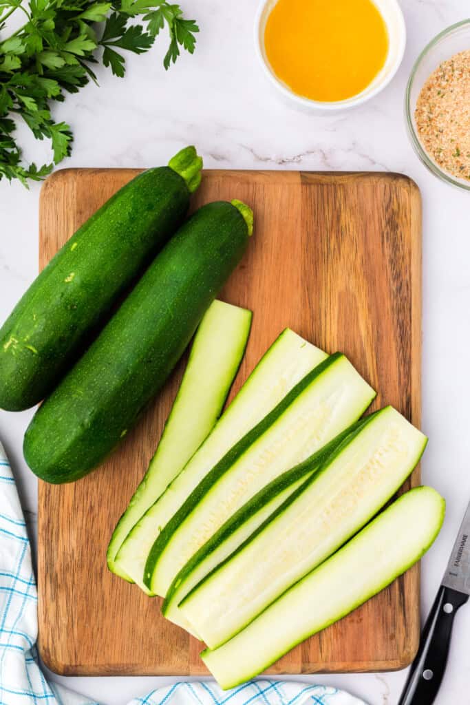 Zucchinis partially sliced into long strips on a wooden cutting board with prep ingredients nearby.