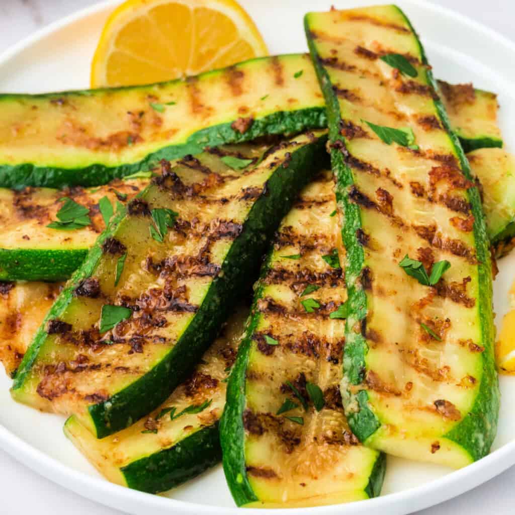 Close-up of grilled zucchini slices with melted parmesan and grill marks, garnished with parsley and lemon.