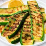 Close-up of grilled zucchini slices with melted parmesan and grill marks, garnished with parsley and lemon.