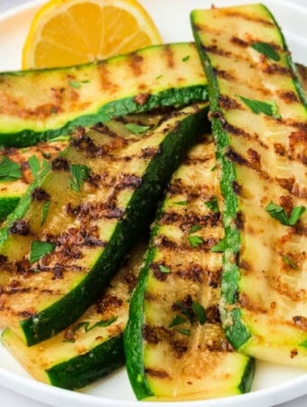 Close-up of grilled zucchini slices with melted parmesan and grill marks, garnished with parsley and lemon.