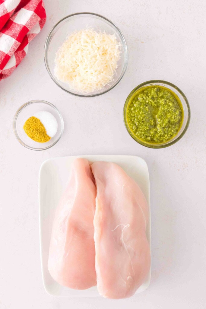Ingredients for grilled pesto chicken: raw chicken breasts, pesto, seasoning, and grated cheese on a light surface.