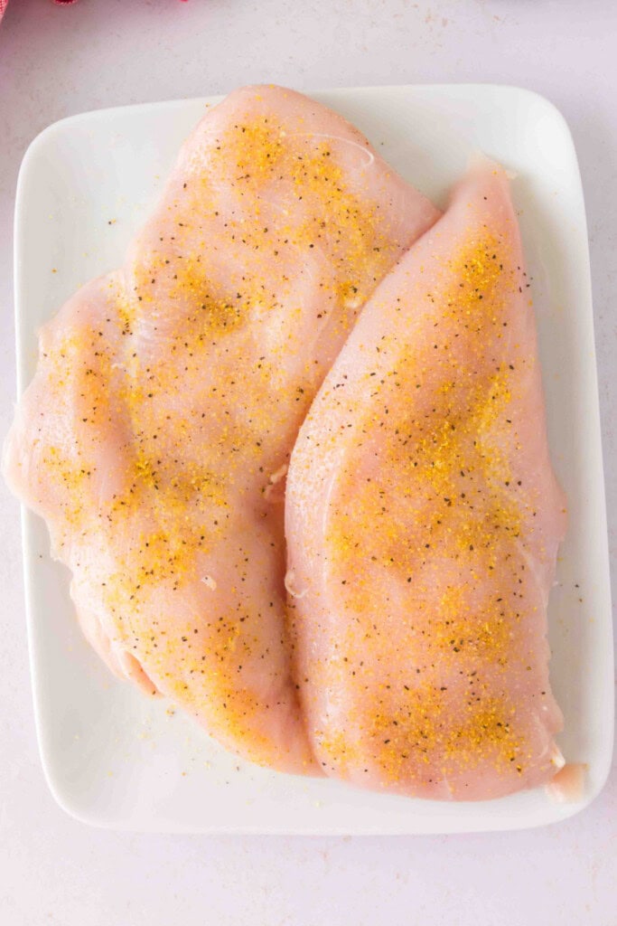 Raw chicken breasts seasoned with salt and lemon pepper on a white plate.