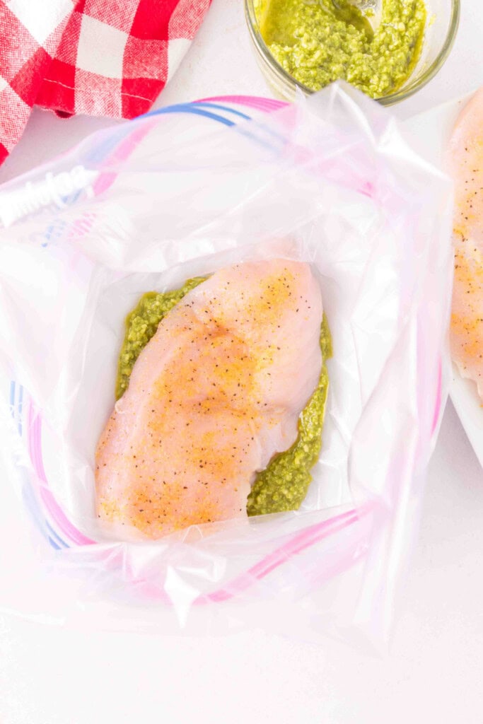 Seasoned chicken breast placed over pesto inside a zip-top bag.