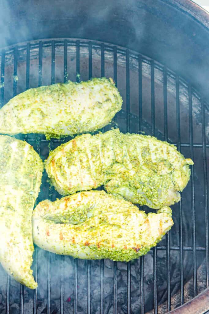 Pesto-coated chicken breasts grilling over open flames with visible smoke.