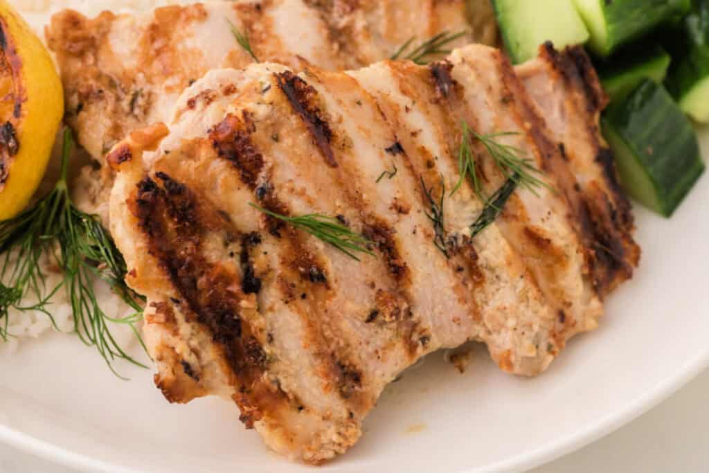 Close-up of grilled tahini chicken thighs served with grilled lemon, fresh dill, and chopped cucumber on a white plate.