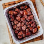 Overhead view of BBQ-glazed Poor Man’s Burnt Ends in a foil pan on a wooden board, square crop.