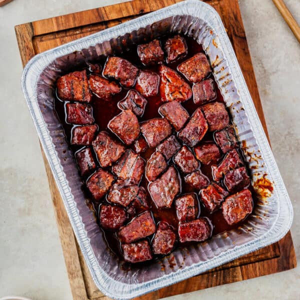 Overhead view of BBQ-glazed Poor Man’s Burnt Ends in a foil pan on a wooden board, square crop.