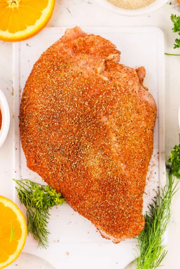 Raw turkey breast coated in Cajun seasoning on a white cutting board surrounded by fresh herbs and orange slices