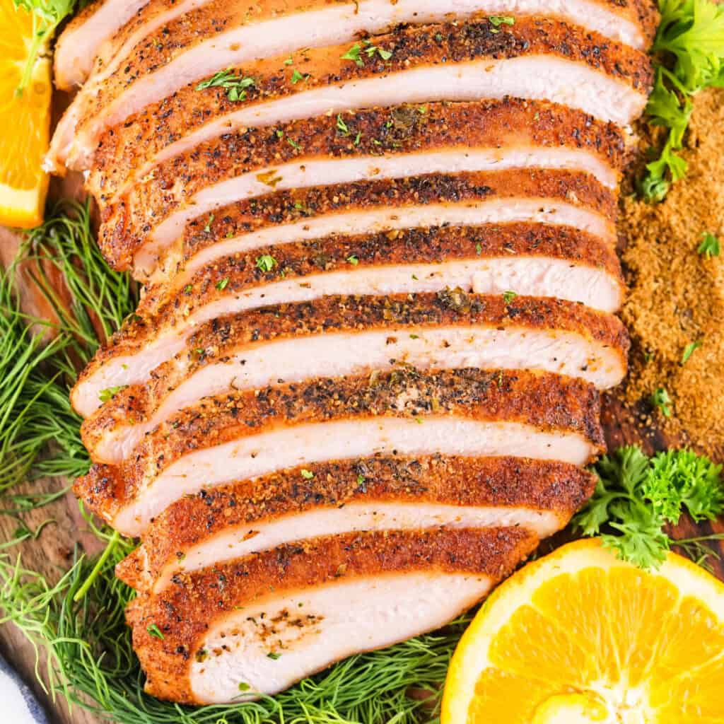 Square image of sliced smoked Cajun turkey breast surrounded by fresh herbs, seasoning, and orange slices