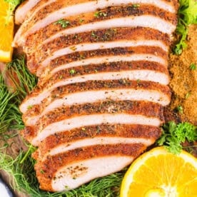 Square image of sliced smoked Cajun turkey breast surrounded by fresh herbs, seasoning, and orange slices