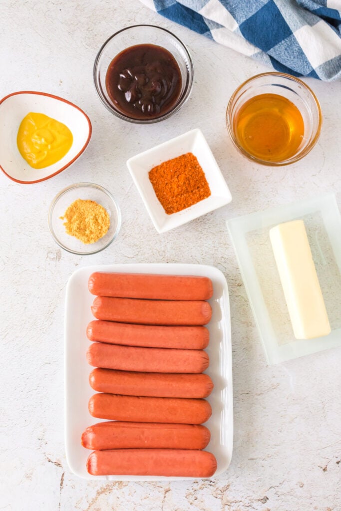 Overhead view of ingredients including hot dogs, butter, mustard, BBQ sauce, honey, and spices on a white surface