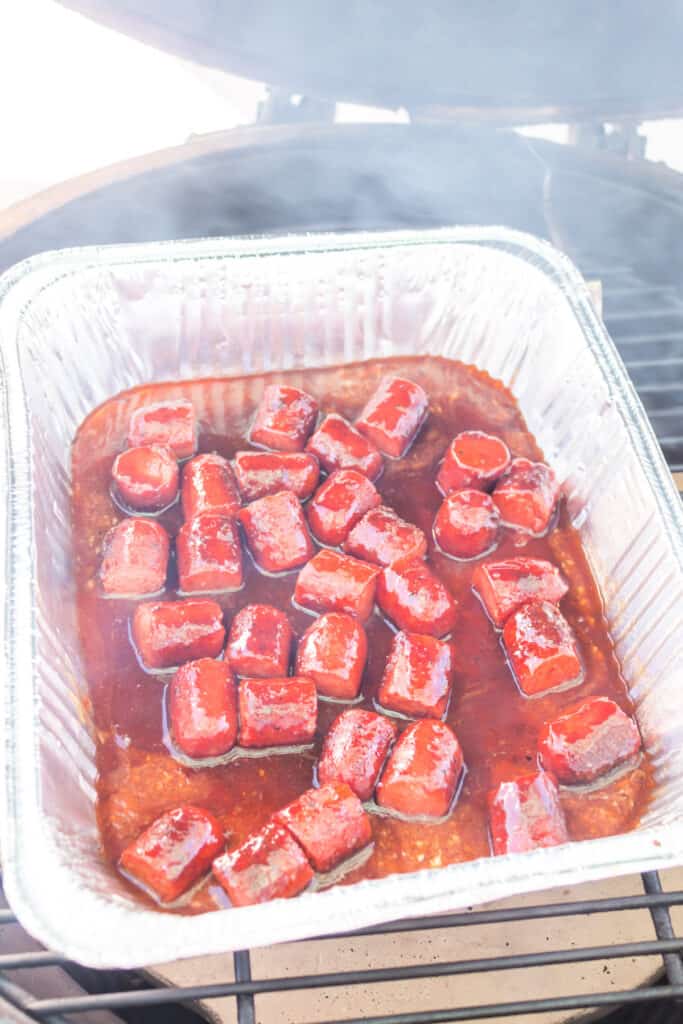 Aluminum tray of saucy hot dog bites sitting on a smoker grill with smoke rising
