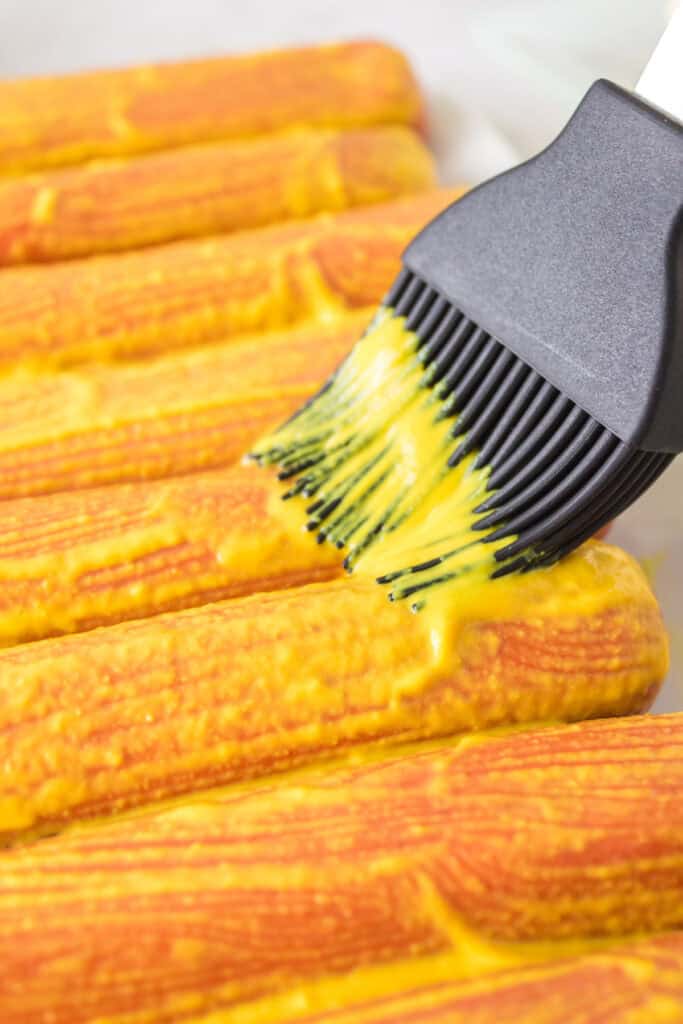 Close-up of a basting brush spreading yellow mustard onto hot dogs before seasoning