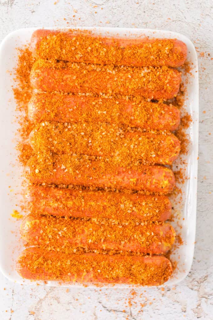 Hot dogs fully coated in dry rub seasoning on a white platter, ready for smoking