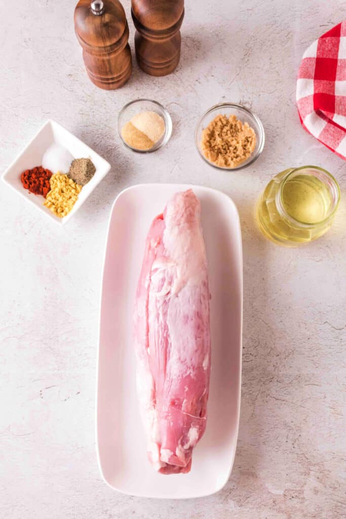 Overhead view of raw pork tenderloin on a white tray surrounded by rub ingredients and olive oil