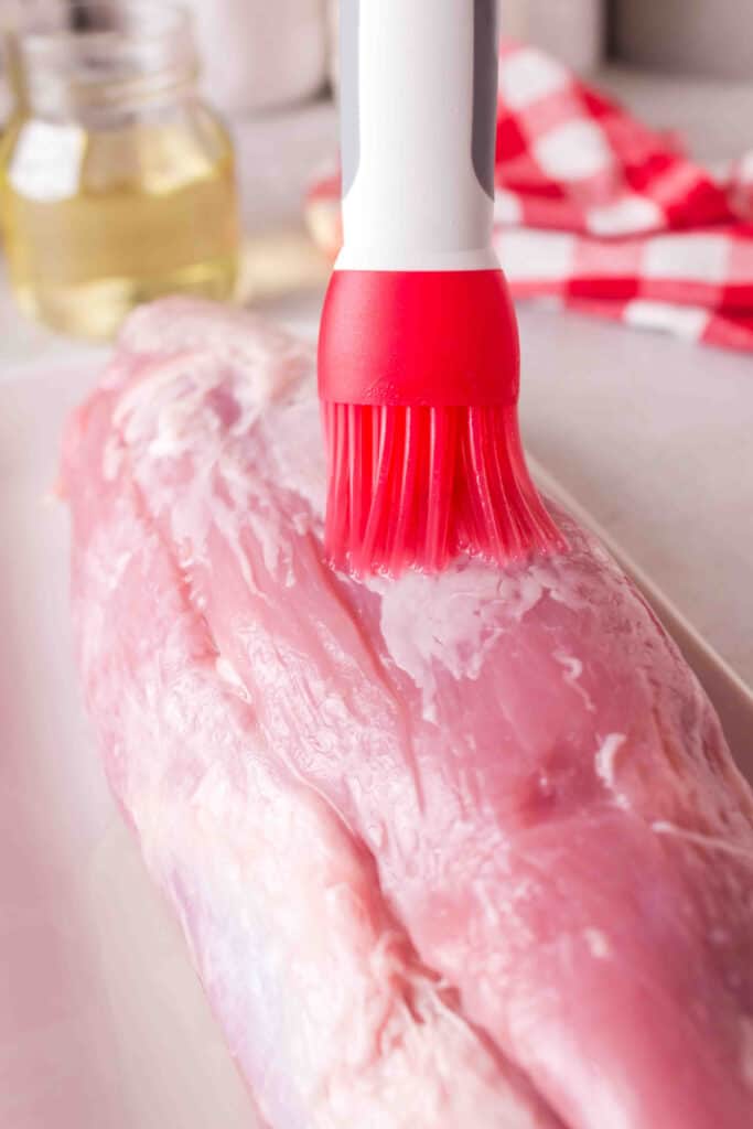 Olive oil being brushed onto raw pork tenderloin using a red silicone brush