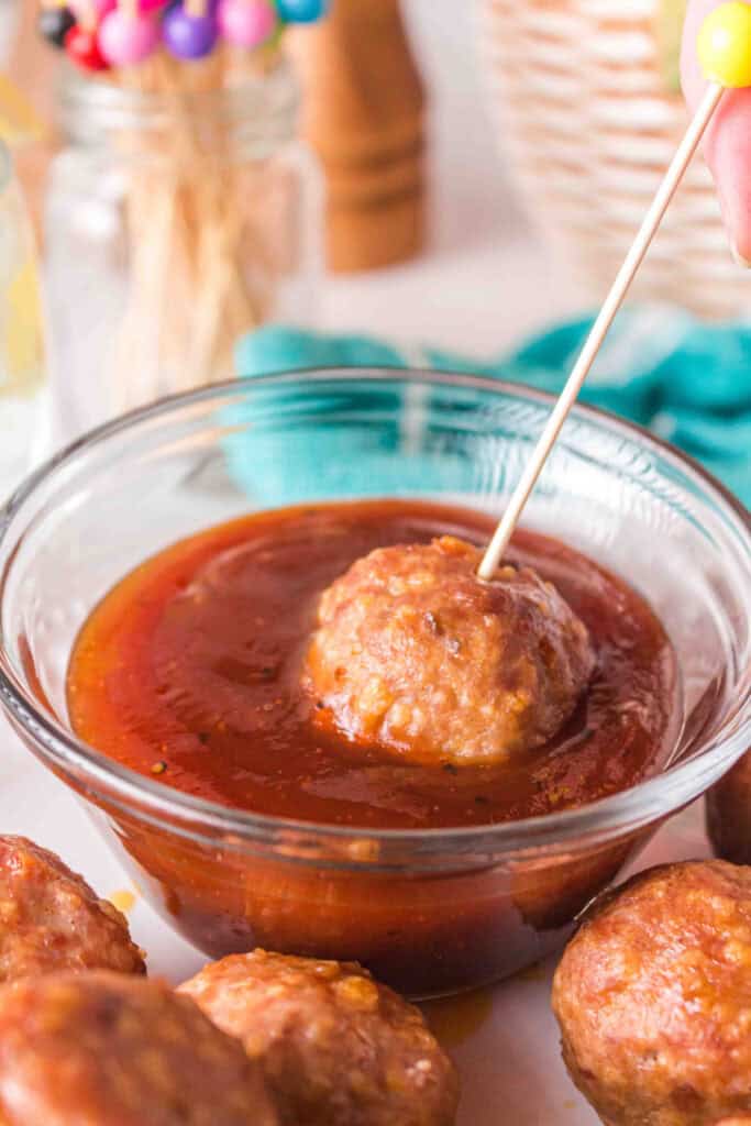 A turkey meatball being dipped into BBQ sauce using a colorful skewer.