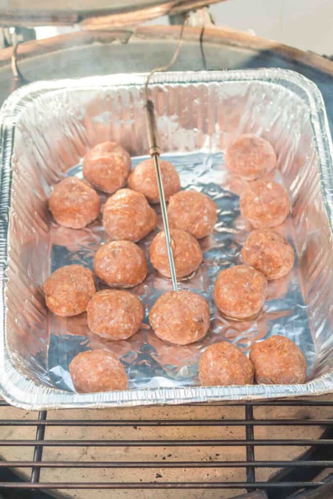Raw turkey meatballs smoking in a foil pan on a grill with a temperature probe.