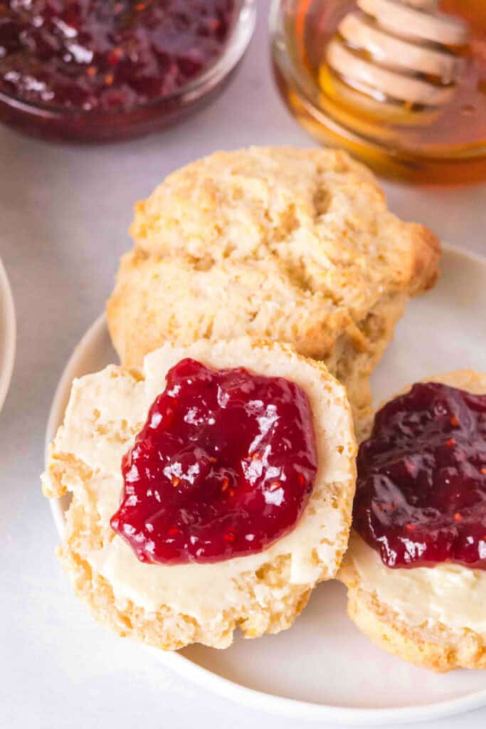 Split scone topped with butter and raspberry jam, with more scones in background