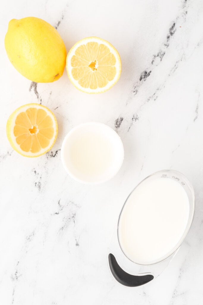 Whole and halved lemons with small bowls of lemon juice and milk on marble.