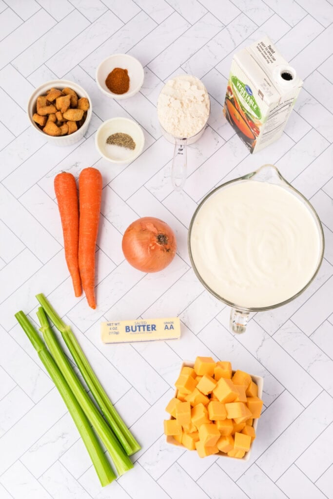 Overhead shot of all cheese soup ingredients laid out on a white tile background, including carrots, celery, onion, Velveeta cubes, butter, flour, and cream.