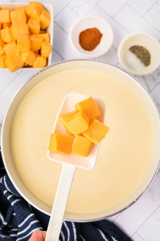 Cubes of Velveeta cheese held on a white spatula above a pot of creamy soup, ready to be stirred in. Bowls of cayenne and pepper nearby.