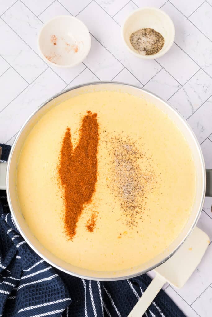 Overhead view of creamy cheese soup with cayenne pepper and ground black pepper sprinkled on top, before stirring.