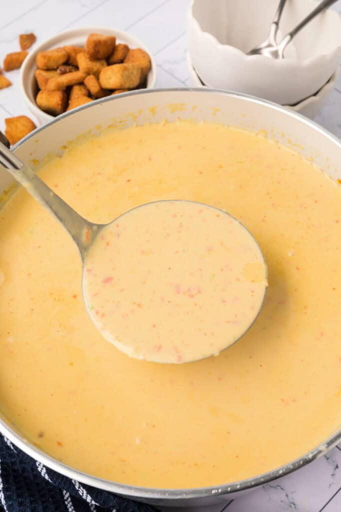 A ladle filled with rich, creamy cheese soup hovering above the pot, with small bits of vegetables visible in the velvety broth.
