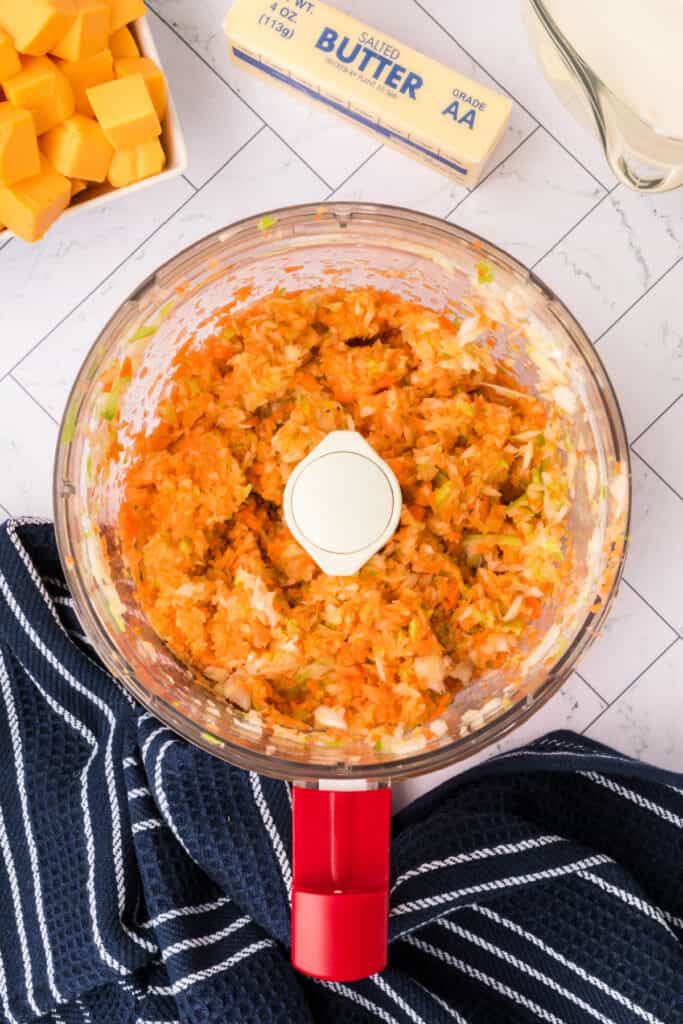 Chopped carrots, onion, and celery blended in a food processor bowl sitting on a navy striped kitchen towel.