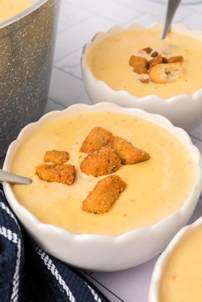 A close-up of a white scalloped bowl filled with creamy cheese soup, topped with golden croutons and served with a spoon.