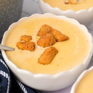 Square-format image showing creamy cheese soup with croutons in a white bowl, placed near a pot and blue striped towel.