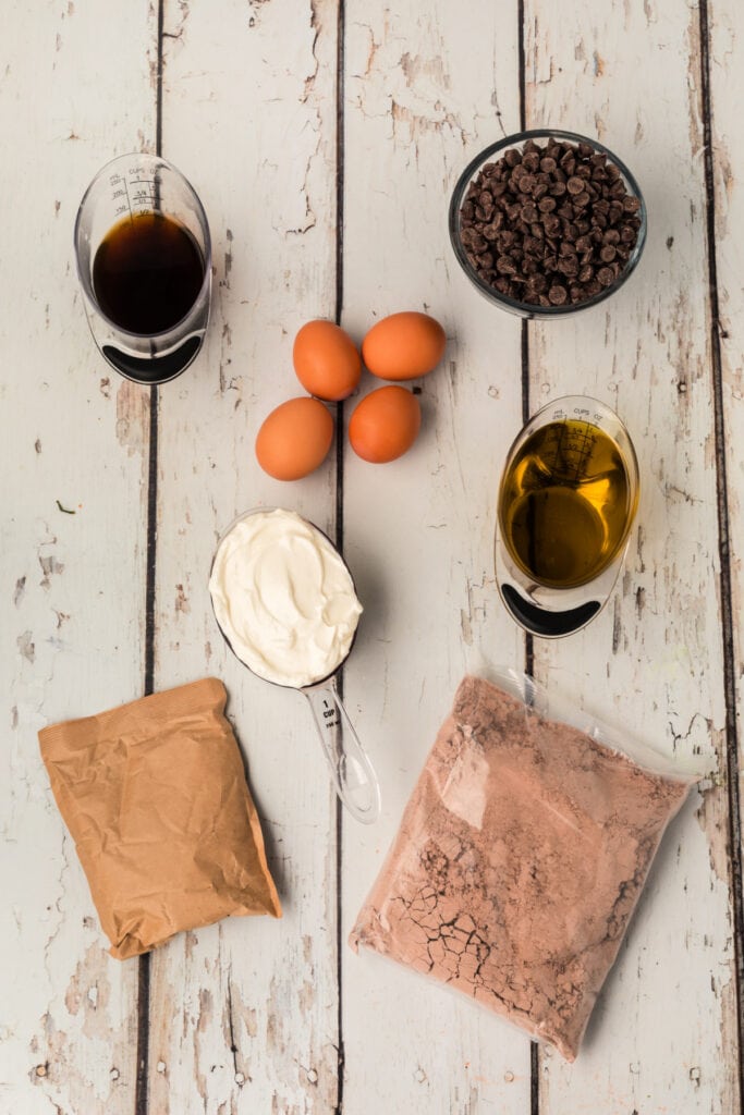 Overhead view of chocolate Bundt cake ingredients including eggs, sour cream, oil, chocolate chips, pudding mix, and coffee on a distressed white wood surface.