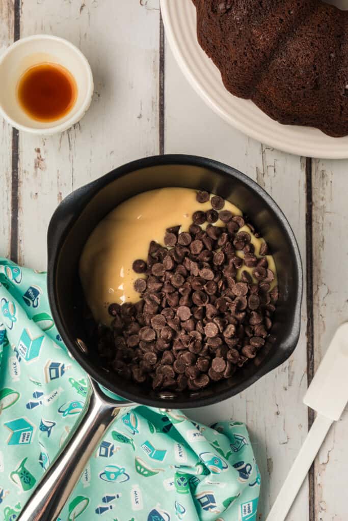 Saucepan filled with sweetened condensed milk and chocolate chips next to a partially visible Bundt cake on a plate.