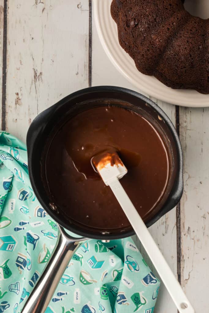 Smooth and glossy chocolate glaze being stirred in a saucepan with a spatula, ready to be poured over the cake.